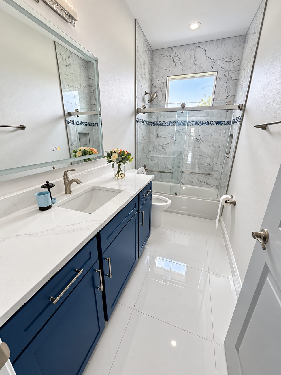 Tub-shower in marble-look porcelain with a blue glass mosaic band, navy vanity and a lit mirror