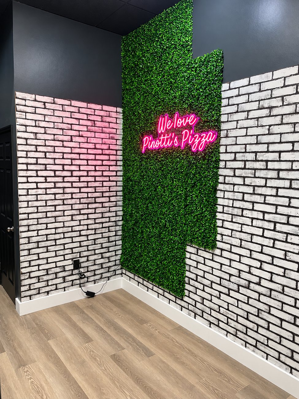 Restaurant corner with white faux brick panel walls, a green foliage wall and a pink neon sign reading We love Pinotti&rsquo;s Pizza