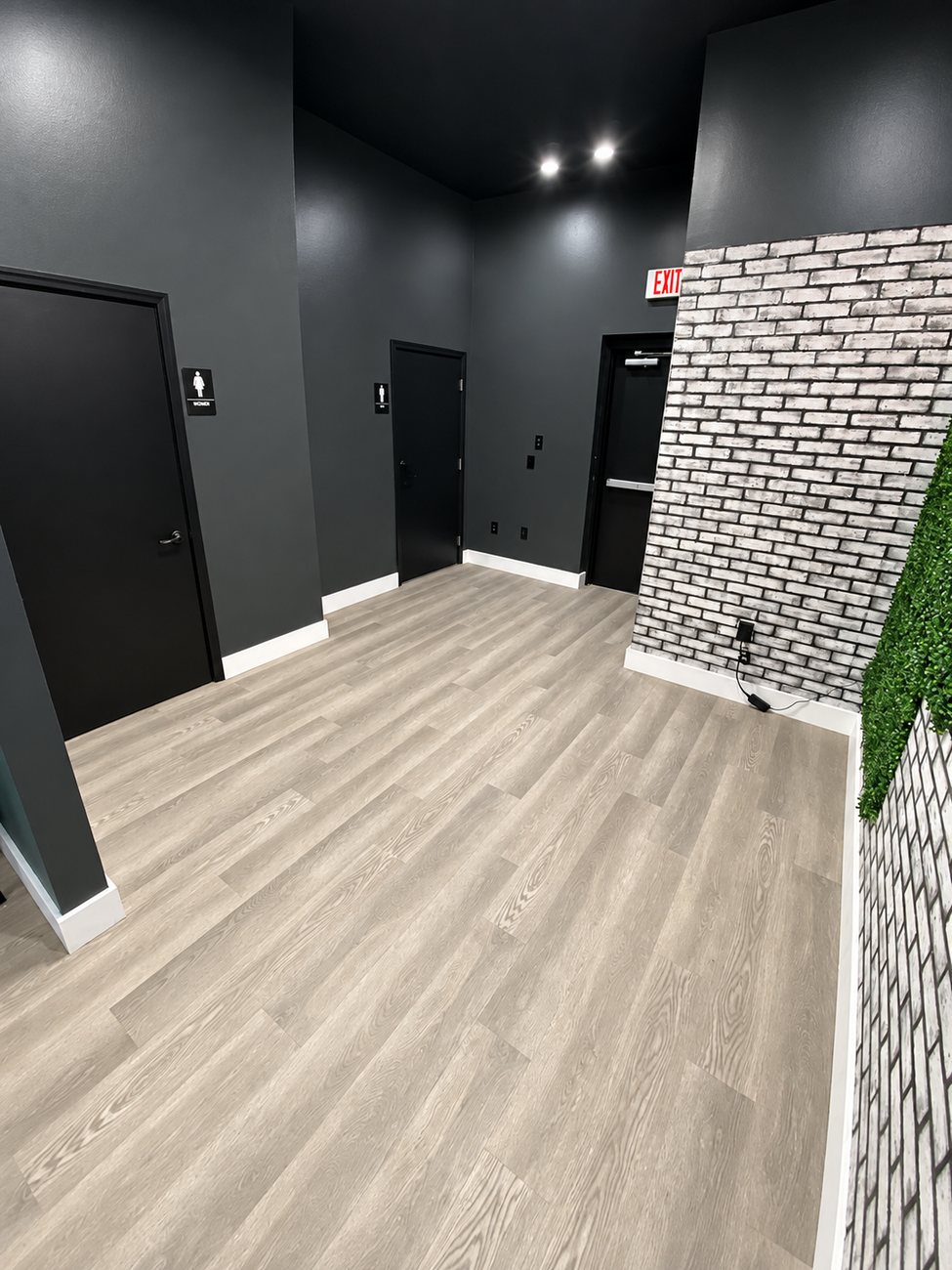 Commercial corridor with charcoal walls, black restroom doors, exit sign and a white brick panel wall over plank flooring
