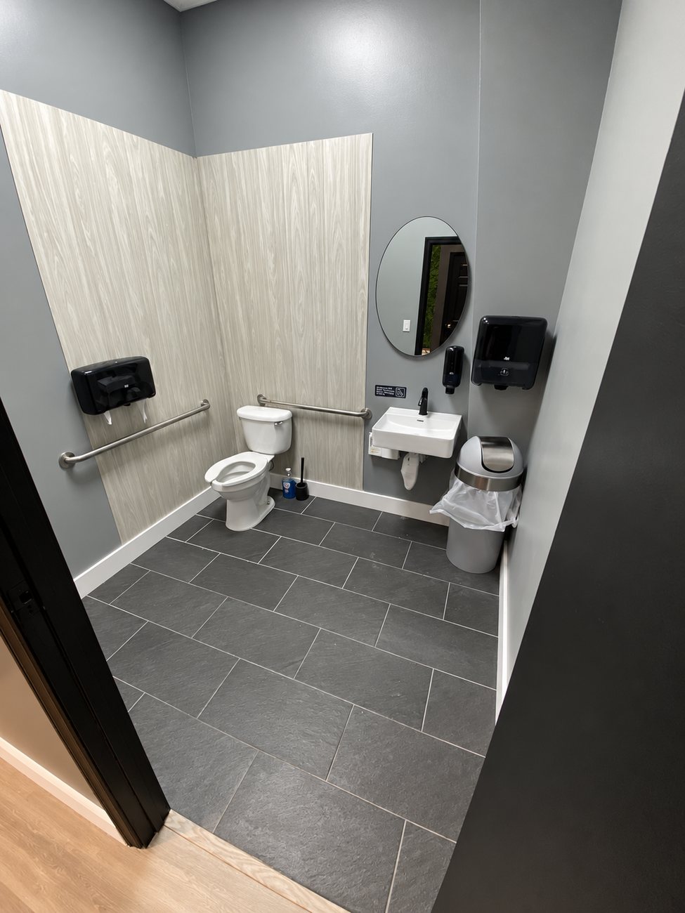 Commercial ADA restroom with grab bars, wall-hung sink, round mirror and dark slate-look tile floor