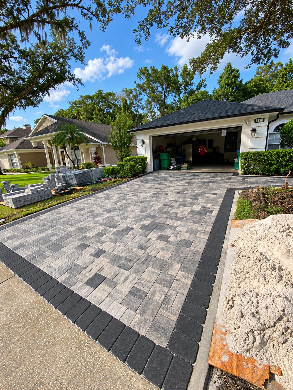 Grey paver driveway with charcoal border under construction, pallets of pavers and a pile of sand on site
